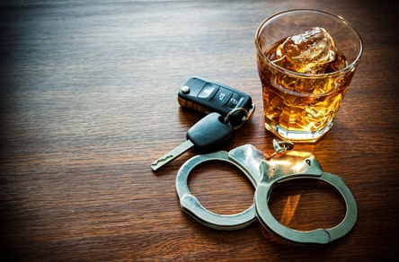 A glass of whiskey with ice, car keys, and handcuffs rest on a wooden table. It symbolizes the serious consequences of impaired driving.