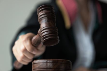 A person in a dark robe holds a wooden gavel, poised to strike a matching sounding block on a table.
