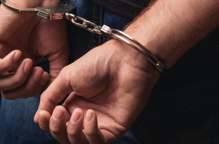 Close-up of a person's hands handcuffed together. The silver handcuffs are prominent against dark clothing, with blue jeans visible.