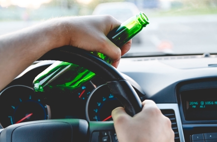 A person's hands are on a car's steering wheel, with one hand holding a green beer bottle. The blurred background suggests the car is in motion.