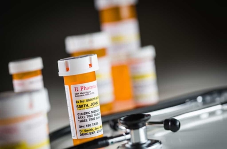 Prescription bottles and a stethoscope. One bottle's label visible: "SMITH, JOHN," "GENERIC MEDICATION," "TAKE TWO TABLETS THREE TIMES TWO DAYS," "QTY: 180 TABS."