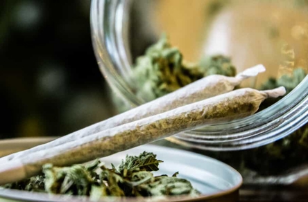A close-up shows two pre-rolled cannabis joints on a surface with loose buds, with a jar of cannabis visible in the background. This image depicts cannabis products.
