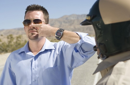 A man in sunglasses performs a field sobriety test, touching his nose as a police officer observes. This image depicts a common roadside interaction related to DWI charges.
