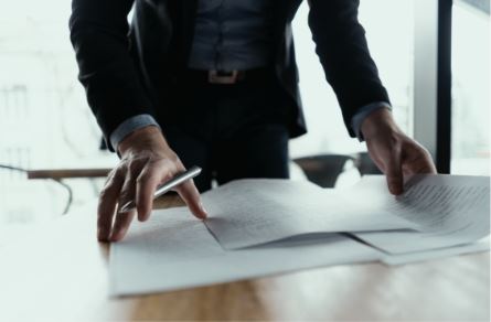 A person in professional attire reviews documents on a table, holding a pen. The scene suggests a focused, administrative task.
