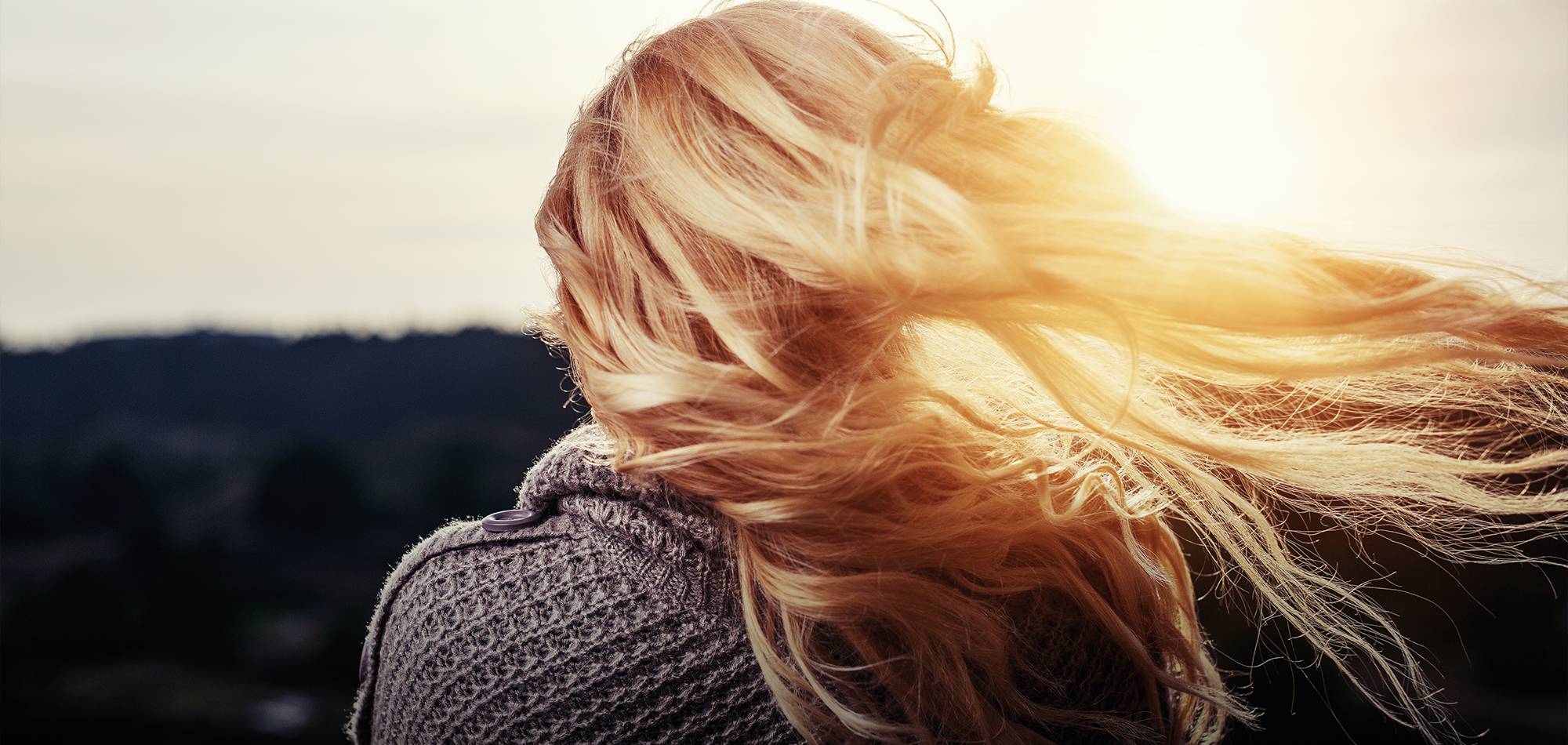 A woman viewed from behind, with long blonde hair flowing in the wind and dramatically backlit by golden sunlight. She wears a gray sweater, looking out over a blurred landscape, suggesting reflection or a hopeful outlook.